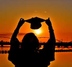 student in cap and gown holding up cap against sunset